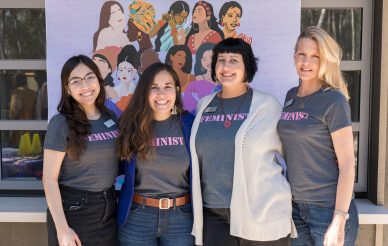 A square comrised of racially diverse women against a light purple background with 4 UCSD staff in the front, all wearinggray t-shirts with the word Feminist emblazoned in pink.